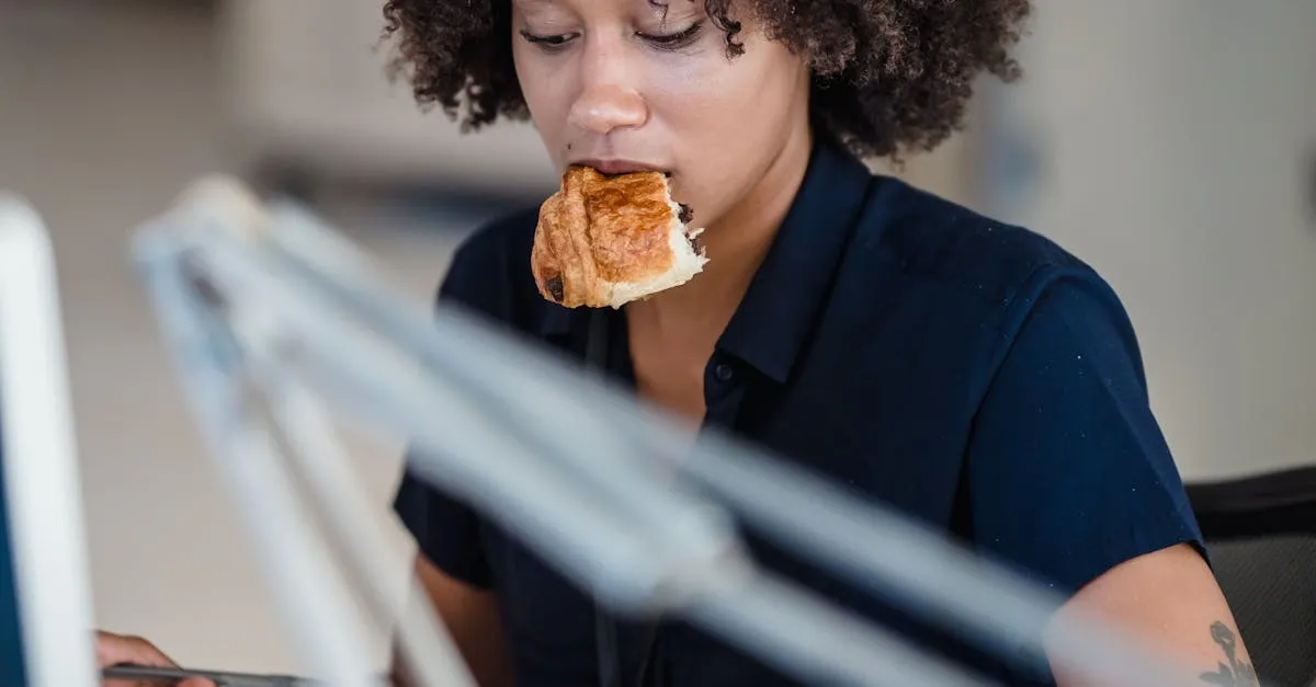 Businesswoman multitasking at her desk, eating a croissant while working on a computer in a busy office.