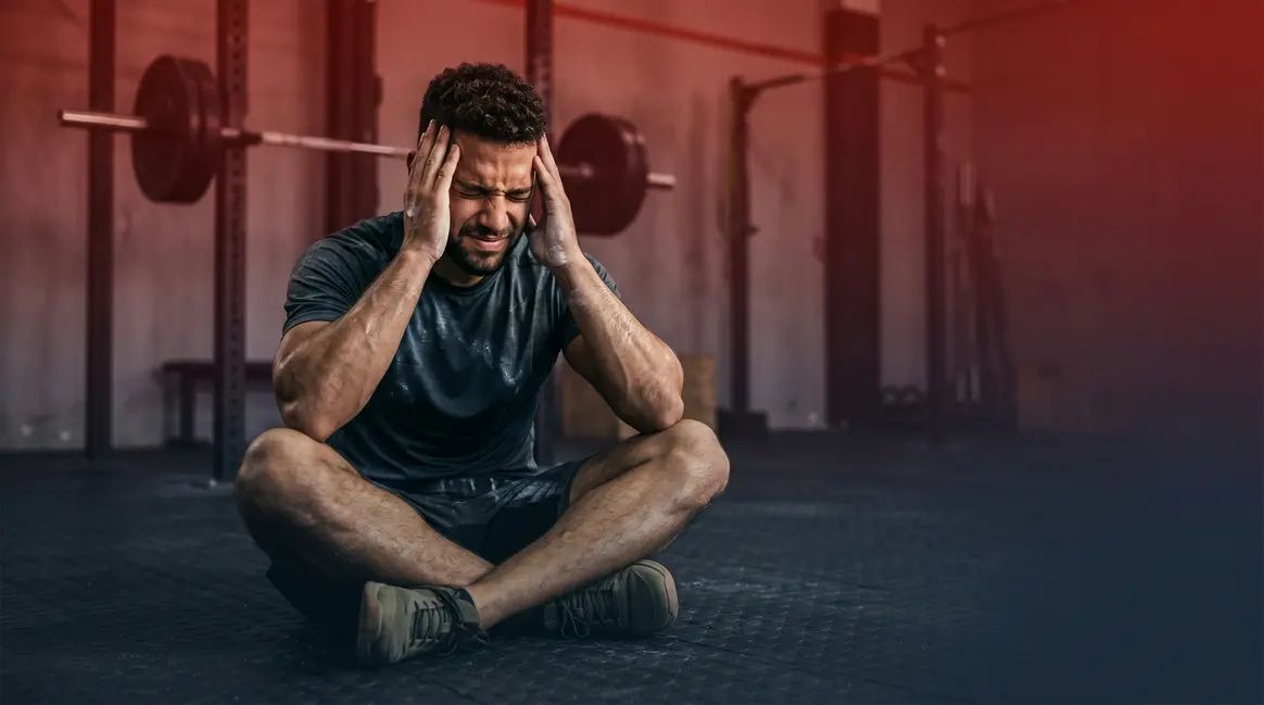 Athlete experiencing headaches after workout sitting on gym floor holding head in pain